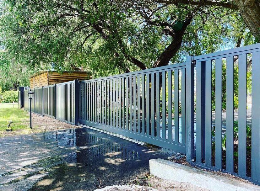 Long metal fence with vertical slats in a garden setting, partially shaded by large trees.